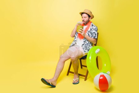 Téléchargez les photos : Beau homme heureux buvant une boisson à l'ananas assis et relaxant avec des inflatables et une balle de plage sur un fond jaune studio - en image libre de droit