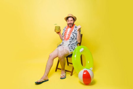 Téléchargez les photos : Joyeux homme heureux portant un short et un chapeau pendant ses vacances d'été en profitant d'une boisson à l'ananas sur un fond jaune - en image libre de droit