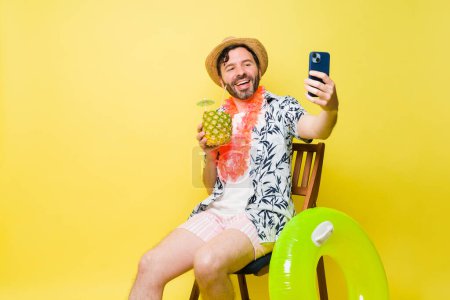 Foto de Feliz hombre hispano sonriendo tomando una selfie para las redes sociales mientras toma una copa en la playa durante un fondo amarillo veraniego - Imagen libre de derechos