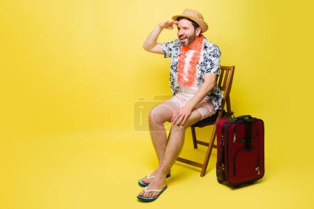 Téléchargez les photos : Souriant homme heureux qui cherche à s'amuser tout en étant prêt à voyager pour les vacances d'été et d'aller à la plage en face d'un fond jaune - en image libre de droit