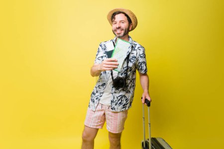 Foto de Alegre hombre caucásico sonriendo sosteniendo su pasaporte y boletos de avión con una maleta lista para viajar a la playa - Imagen libre de derechos