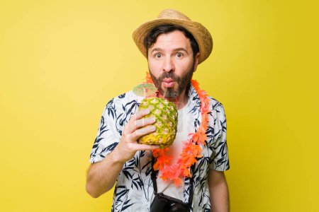 Téléchargez les photos : Homme attrayant dans sa quarantaine avec un chapeau boire une boisson à l'ananas profiter de voyager à la plage pour des vacances d'été sur un fond jaune - en image libre de droit