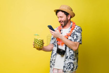 Téléchargez les photos : Un touriste joyeux prenant une photo de sa boisson à l'ananas tout en profitant de ses vacances d'été à la plage sur fond jaune - en image libre de droit
