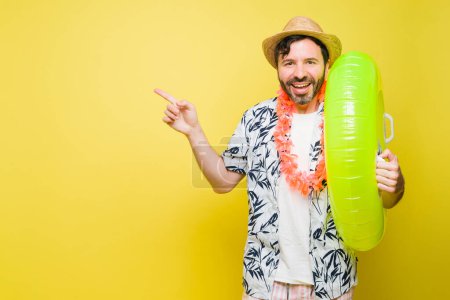 Téléchargez les photos : Latin homme dans son 40s rire et pointant vers copier l'espace en face d'un fond jaune tout en appréciant la plage et la piscine pendant les vacances - en image libre de droit