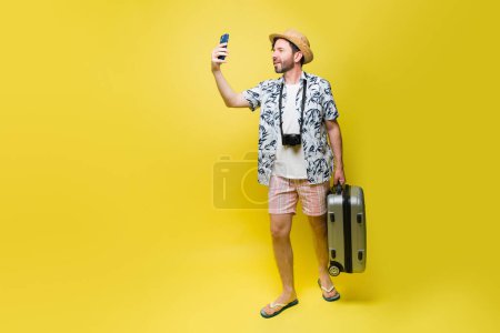 Téléchargez les photos : Heureux touriste caucasien avec un short et un chapeau prendre des photos et selfies avec son téléphone tout en portant une valise à ses vacances d'été - en image libre de droit