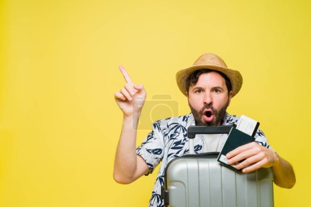 Foto de Sorprendido hombre adulto medio mirando emocionado mientras señala hacia arriba con su dedo y listo para viajar en las vacaciones de verano delante de un fondo amarillo - Imagen libre de derechos