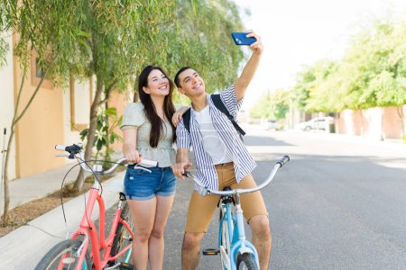 Photo for Attractive couple taking a selfie with a smartphone while smiling together and having a bike ride outdoors during a beautiful summer day - Royalty Free Image