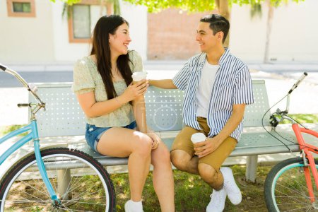 Photo for Attractive relaxed happy couple talking on the bench after going on a bike ride in the park and drinking coffee outdoors during a date - Royalty Free Image