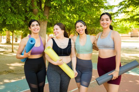 Téléchargez les photos : Souriant belles femmes diversifiées avec des tapis de yoga regardant heureux dans les vêtements de sport prêts à commencer leurs exercices en plein air pour l'acceptation du corps - en image libre de droit