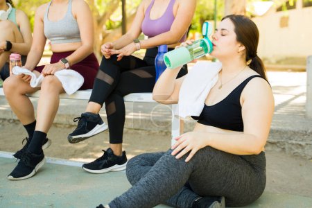 Photo for Plus size young woman drinking water after sweating from workout exercises or run outdoors with a group of diverse women - Royalty Free Image
