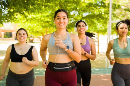 Foto de Alegre joven latina sonriendo haciendo contacto visual mientras corre por el parque y hace ejercicio con un grupo diverso de amigas - Imagen libre de derechos