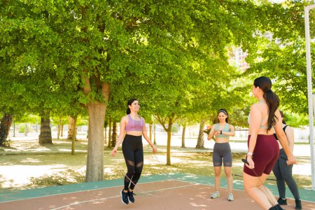 Téléchargez les photos : Groupe heureux de diverses femmes souriant corde sautante et l'exercice au cours d'un entraînement cardio à l'extérieur - en image libre de droit