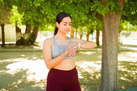 Téléchargez les photos : Jeune femme sportive utilisant une application de fitness sur sa smartwatch avant de commencer à courir ou ses exercices dans le parc - en image libre de droit