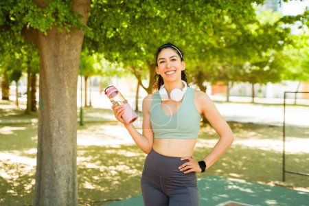Photo for Fit active woman looking sporty in the park wearing activewear smiling drinking water after running or doing cardio exercises - Royalty Free Image