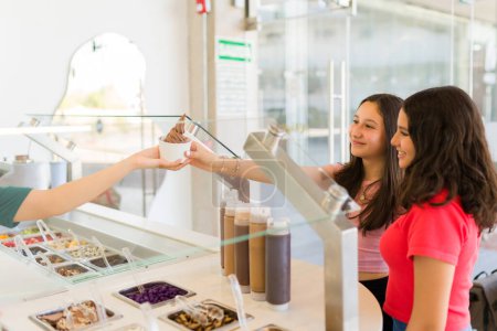 Foto de Chicas adolescentes felices comprando delicioso helado con ingredientes para el postre y sonriendo mientras están en la tienda de yogur congelado - Imagen libre de derechos