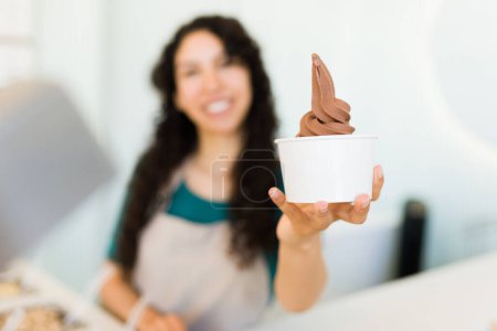 Photo for Focus on foreground of a happy woman worker selling delicious chocolate ice cream or gelato at the frozen yogurt store - Royalty Free Image