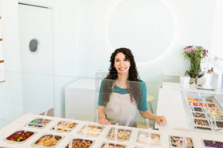 Téléchargez les photos : Jolie travailleuse latine avec un tablier au comptoir du bar souriant tout en travaillant au magasin de yaourts glacés ou à la boutique de crème glacée - en image libre de droit
