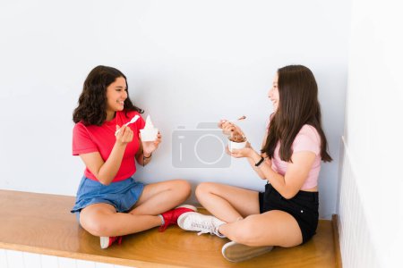 Foto de Adolescentes hispanas hablando de chismes y sonriendo felices mientras comen delicioso helado o yogur congelado - Imagen libre de derechos