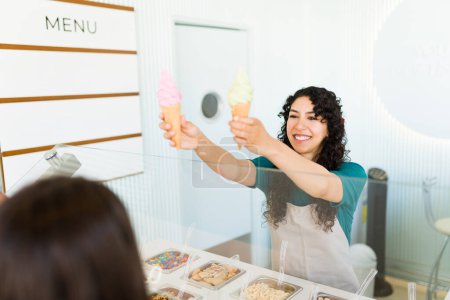 Photo for Cheerful happy woman worker smiling while selling two ice cream cones to customers at the frozen yogurt shop - Royalty Free Image