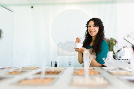 Photo for Beautiful happy woman worker smiling looking at the camera while eating delicious frozen yogurt or ice cream behind the toppings bar - Royalty Free Image