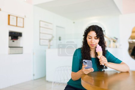 Téléchargez les photos : Jeune femme hispanique texter sur le smartphone tout en mangeant de délicieux cône de crème glacée à la boutique de yaourts glacés - en image libre de droit