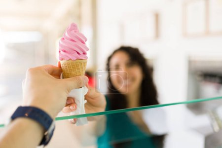 Photo for Woman worker selling a delicious cold strawberry ice cream cone to a happy female customer behind the counter at the gelato shop - Royalty Free Image