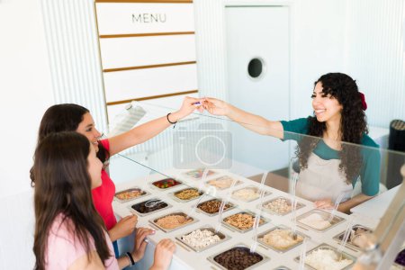 Téléchargez les photos : Travailleur femme heureuse donnant gratuitement des échantillons de crème glacée ou de yaourt congelé pour essayer pour les clients adolescents à la boutique de crème glacée - en image libre de droit