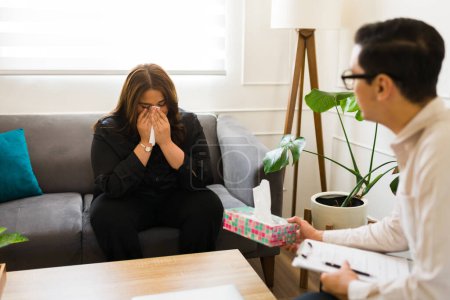 Foto de Triste mujer de luto sonándose la nariz mientras lloraba hablando con un psicólogo profesional sobre su depresión y dolor - Imagen libre de derechos