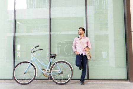 Photo for Happy relaxed young man enjoying the city after commuting to work in his eco friendly bicycle leaning on the urban building - Royalty Free Image
