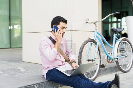 Téléchargez les photos : Homme d'affaires faisant un appel d'affaires à l'extérieur de son bâtiment de travail après avoir fait la navette sur son vélo dans la ville - en image libre de droit