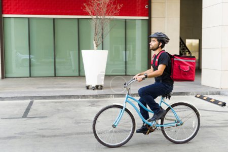 Foto de Vista lateral de un trabajador masculino con un casco que lleva una mochila y montar en bicicleta trabajando en la entrega de alimentos en la ciudad - Imagen libre de derechos