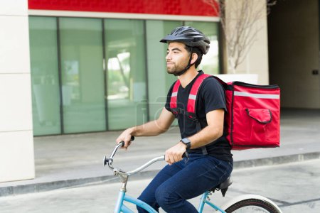 Foto de Atractiva latina montando en bicicleta y trabajando en comida a domicilio mientras lleva una mochila y un casco de seguridad en la ciudad - Imagen libre de derechos