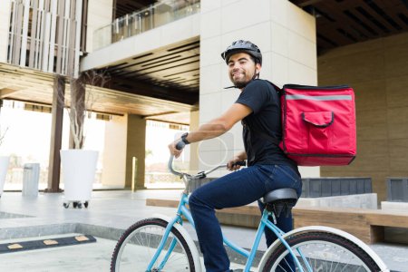 Foto de Joven latino sonriendo y montando en bicicleta en la ciudad mientras trabaja para un servicio de comida a domicilio luciendo feliz - Imagen libre de derechos