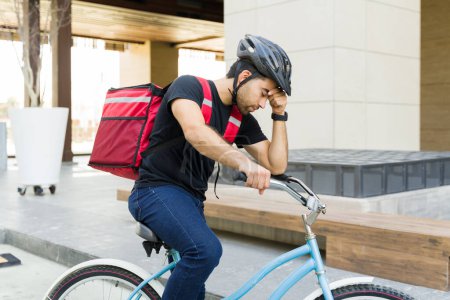Foto de Trabajador de reparto de comida cansado que parece agotado y preocupado mientras trabaja en bicicleta en la ciudad - Imagen libre de derechos