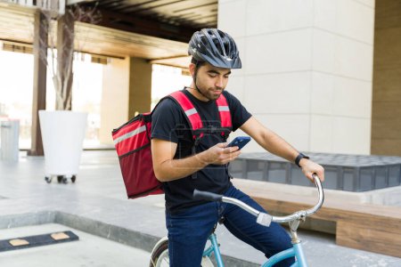 Foto de Joven mirando la aplicación de entrega en su teléfono inteligente mientras espera para hacer una entrega de comida en bicicleta - Imagen libre de derechos