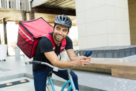 Foto de Alegre trabajador latino sonriendo mientras monta su bicicleta y trabaja en el servicio de comida a domicilio llevando una mochila - Imagen libre de derechos