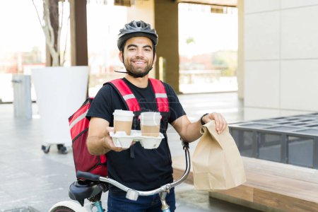 Foto de Emocionado hombre atractivo entregando café y comida almuerzo mientras sonríe y trabaja en el servicio de entrega en bicicleta - Imagen libre de derechos