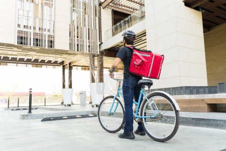 Foto de Joven y repartidor trabajando entregando comida y haciendo mandados vistos desde atrás montando una bicicleta en la ciudad - Imagen libre de derechos