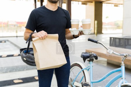 Foto de Primer plano de un joven llevando café y una bolsa de almuerzo mientras trabajaba para una aplicación de entrega de comida usando una bicicleta en la ciudad - Imagen libre de derechos