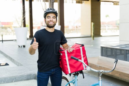 Téléchargez les photos : Jeune homme hispanique faire un pouce vers le haut et à la recherche heureux de travailler la livraison de nourriture dans la ville pour un service d'application de livraison en vélo - en image libre de droit