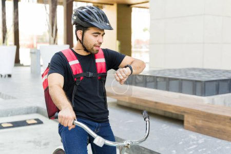 Foto de Trabajador de reparto guapo mirando su reloj y sintiéndose tarde mientras monta su bicicleta y entrega de alimentos - Imagen libre de derechos
