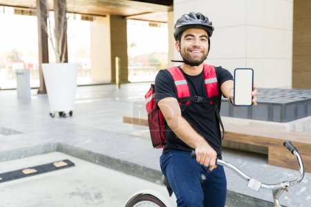 Foto de Emocionado joven que trabaja como repartidor y muestra la pantalla de su smartphone mientras usa la aplicación de comida a domicilio y monta en bicicleta - Imagen libre de derechos