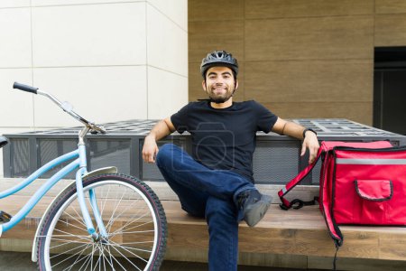 Foto de Trabajador de reparto relajado descansando y sentado en la ciudad esperando para hacer una entrega con una mochila y su bicicleta ecológica - Imagen libre de derechos