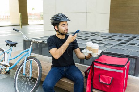 Foto de Joven latino tomando una foto con su smartphone al café mientras hace una entrega de comida y trabaja usando una bicicleta - Imagen libre de derechos