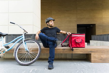 Foto de Joven sonriente descansando y sonriendo mientras trabaja como repartidor montando en bicicleta y llevando una mochila roja - Imagen libre de derechos