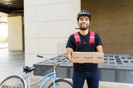 Foto de Alegre repartidor llevando pizzas y sonriendo mientras reparte comida en la ciudad montando en bicicleta - Imagen libre de derechos