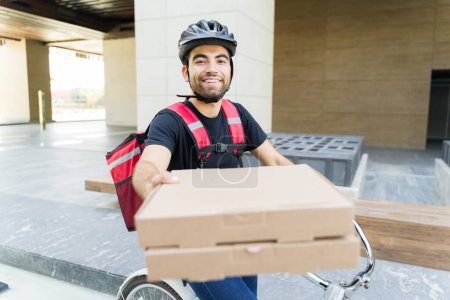 Foto de Feliz hombre latino entregando pizza y mientras trabajaba para una aplicación de comida a domicilio montando una bicicleta y sonriendo - Imagen libre de derechos