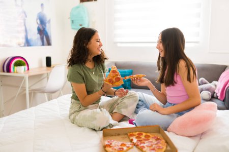 Téléchargez les photos : Joyeux heureux adolescent amis rire tout en appréciant manger pizza ensemble pendant les loisirs détente dans la chambre - en image libre de droit