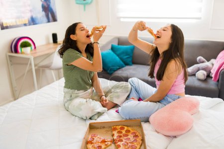 Foto de Feliz adolescentes disfrutando comiendo deliciosa pizza juntos en casa mientras se divierten pasando el rato en la cama - Imagen libre de derechos