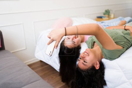 Photo for Smiling teen best friends relaxing in bed together while hanging out and texting on their phone or using social media - Royalty Free Image